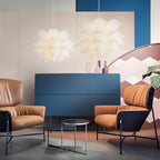 Two Clérachic white petal-layered pendants glowing above a navy sideboard and tan leather armchairs in a colourful room.