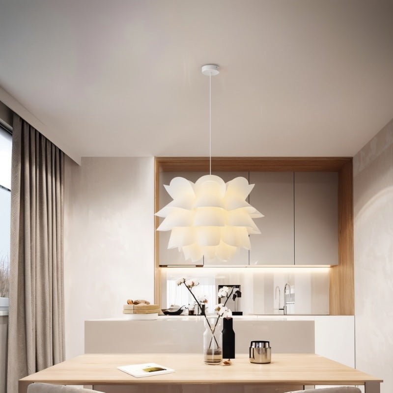 Clérachic white petal-layered pendant glowing over a light wood dining table in front of oak-framed kitchen cabinets.