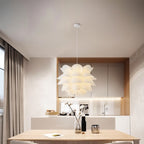 Clérachic white petal-layered pendant glowing over a light wood dining table in front of oak-framed kitchen cabinets.