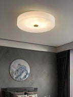 The Clérachic drum ceiling light glowing warm over a grey bedroom, sitting flush to the ceiling above a framed landscape roundel.