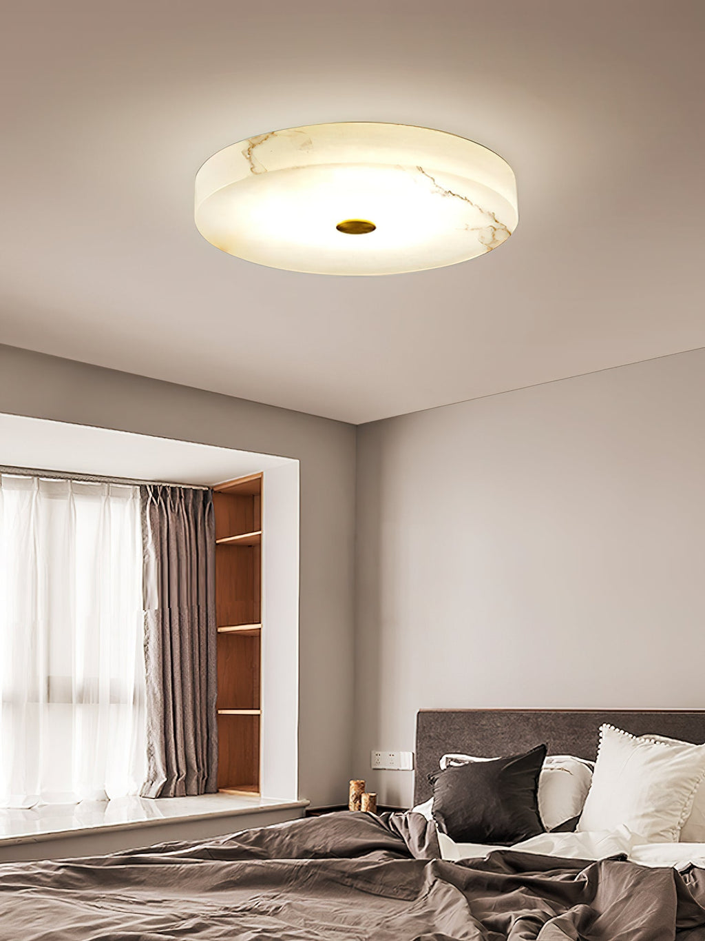 Clérachic alabaster flush mount ceiling light above a bedroom bay window, its stone veining rising warm through the lit surface.