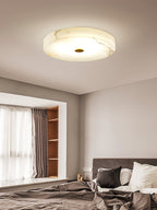 Clérachic alabaster flush mount ceiling light above a bedroom bay window, its stone veining rising warm through the lit surface.