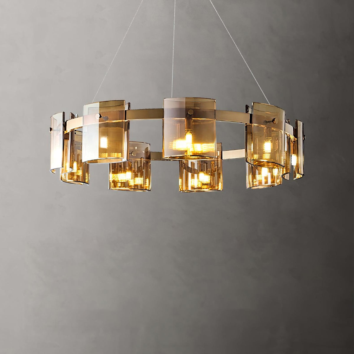 Gold-frame chandelier from Clérachic with amber glass shades lit, shown close on a grey backdrop with its thin hanging cables.