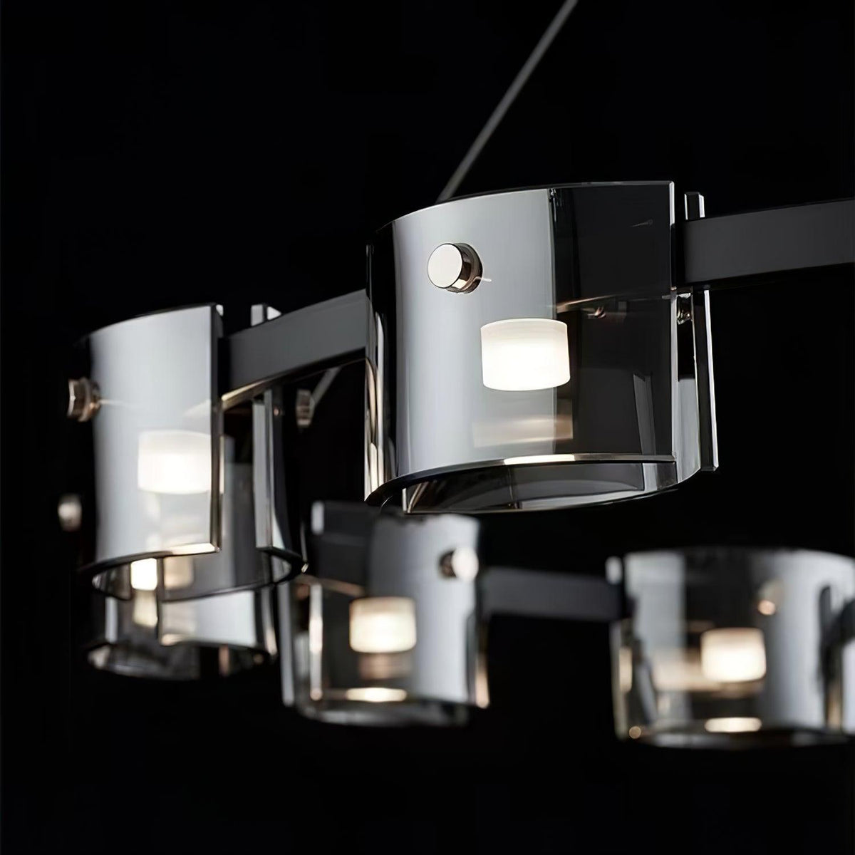 Close view of smoked glass shades on this Clérachic chandelier, with a polished metal pin on each and a bulb glowing within, on a black backdrop.