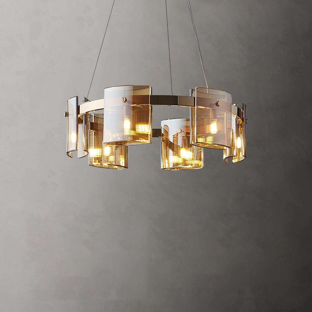 Gold-frame ring chandelier from Clérachic with amber glass shades, lit warm and hung on thin cables against a grey wall.