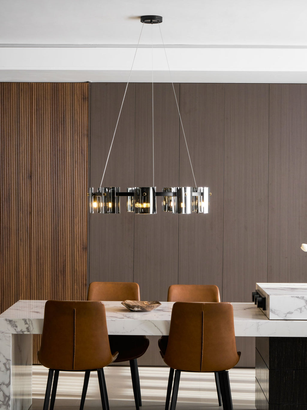 Black-frame Clérachic chandelier hung low above a marble dining table, with tan leather chairs and a ribbed wood wall.