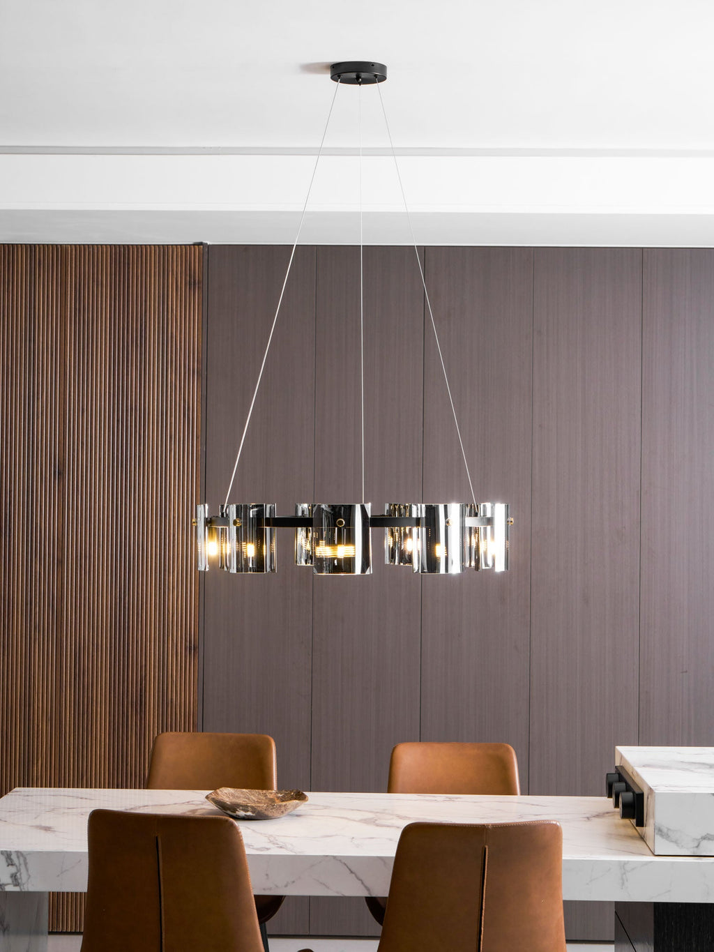 Lit black ring chandelier from Clérachic hung in a dining room with a ribbed wood panel wall and a marble table.