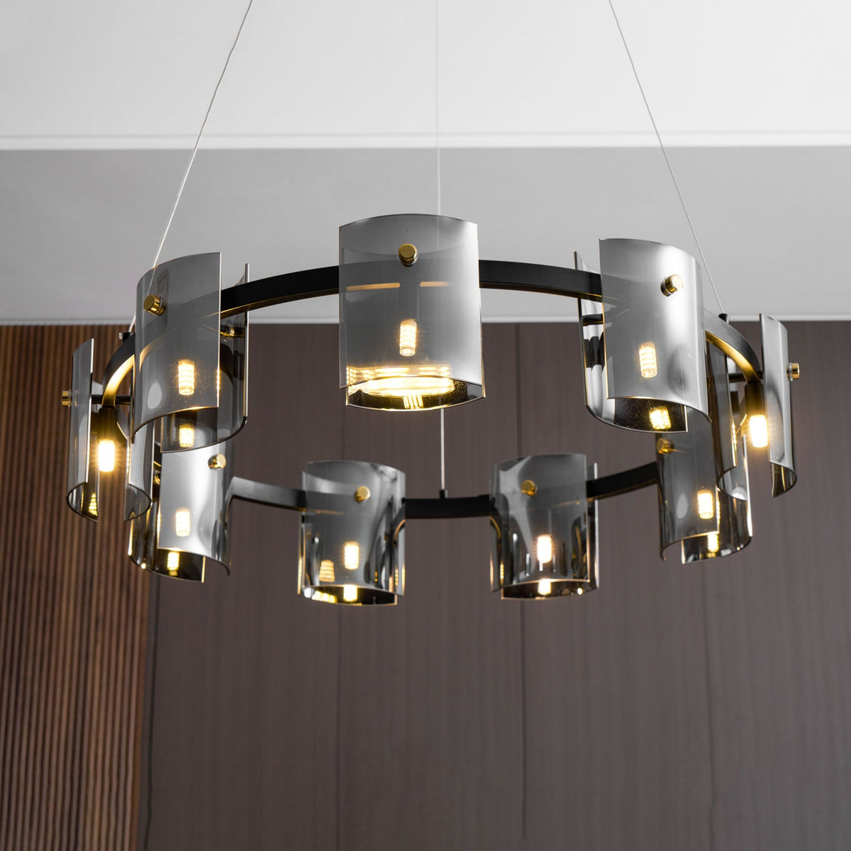 Lit black ring chandelier from Clérachic seen from below, with smoked glass cylinders and brass-coloured pins on a white ceiling.