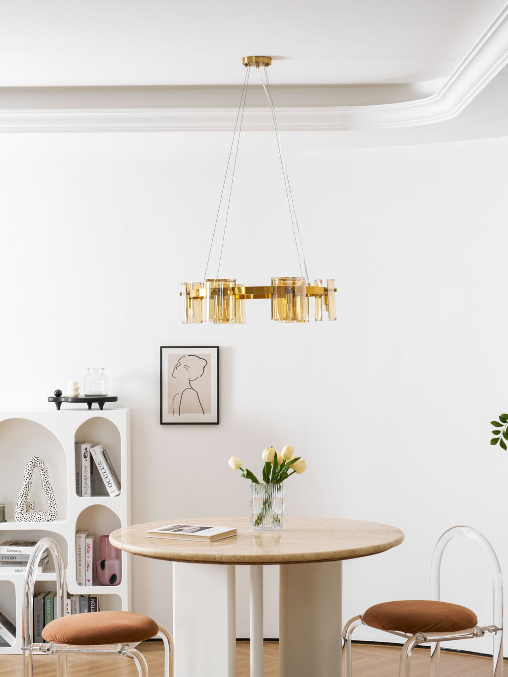 Gold-frame Clérachic chandelier with amber glass above a round travertine table, with arched white shelving behind.