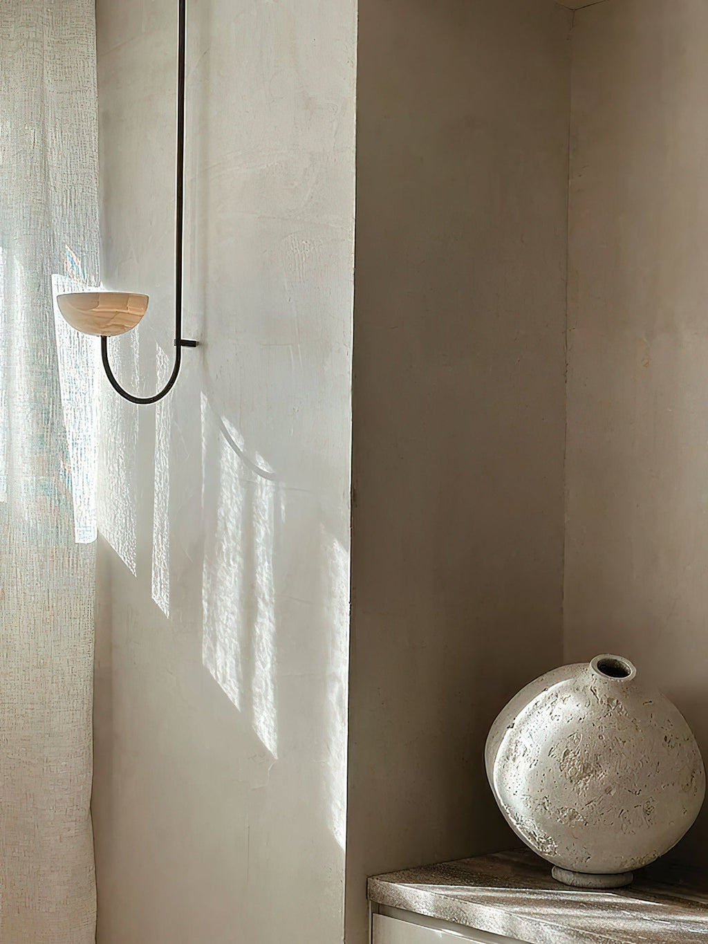 The Clérachic Lunaris single light on plaster beside a linen curtain and a stone vase.
