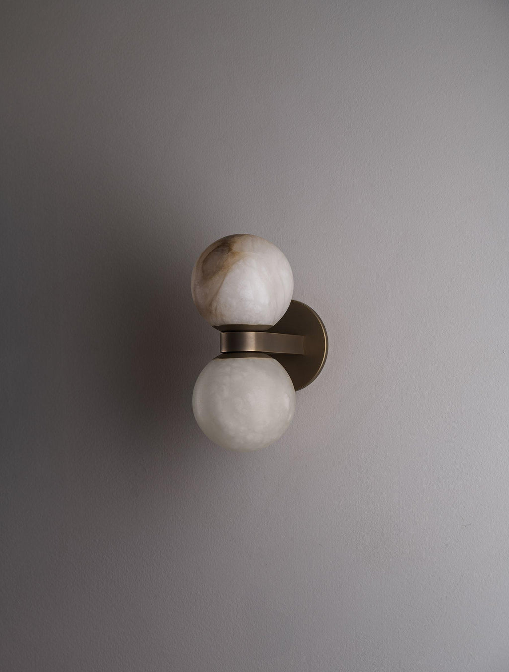 The Clérachic Lush Twin Globe Wall Sconce unlit on a pale plaster wall, its bronze arm and stacked alabaster globes reading as a quiet detail.