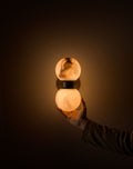 A hand cupping the lower globe of the Clérachic Lush Twin Globe Wall Sconce in a dark room, showing how small the fixture really is.