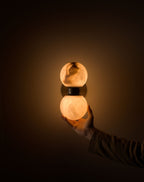 A hand cupping the lower globe of the Clérachic Lush Twin Globe Wall Sconce in a dark room, showing how small the fixture really is.