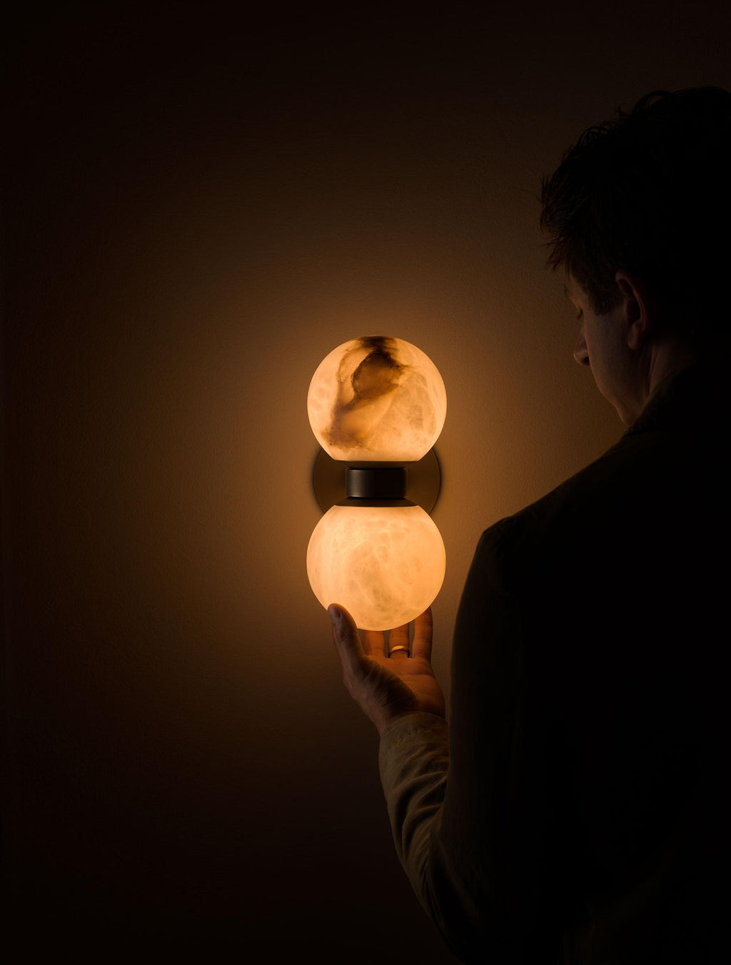 Someone reaching for the glowing lower globe of the Clérachic Lush Twin Globe Wall Sconce on a bedroom wall in an unlit room.