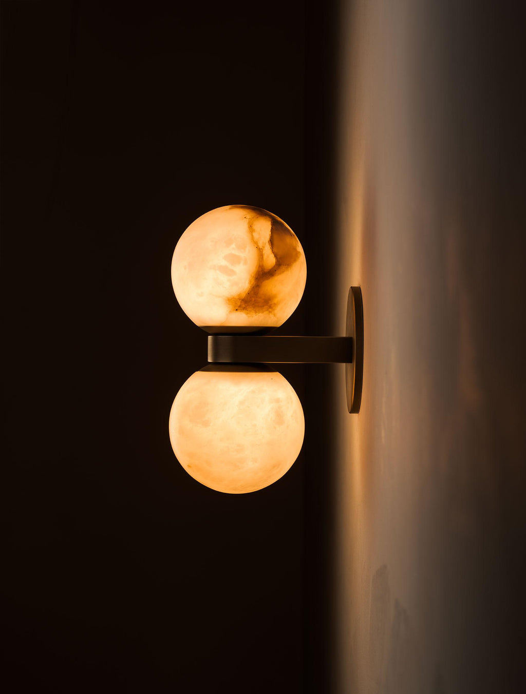 The Clérachic Lush Twin Globe Wall Sconce seen from the side and lit, warm light washing the wall behind its bronze backplate.