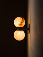 The Clérachic Lush Twin Globe Wall Sconce seen from the side and lit, warm light washing the wall behind its bronze backplate.