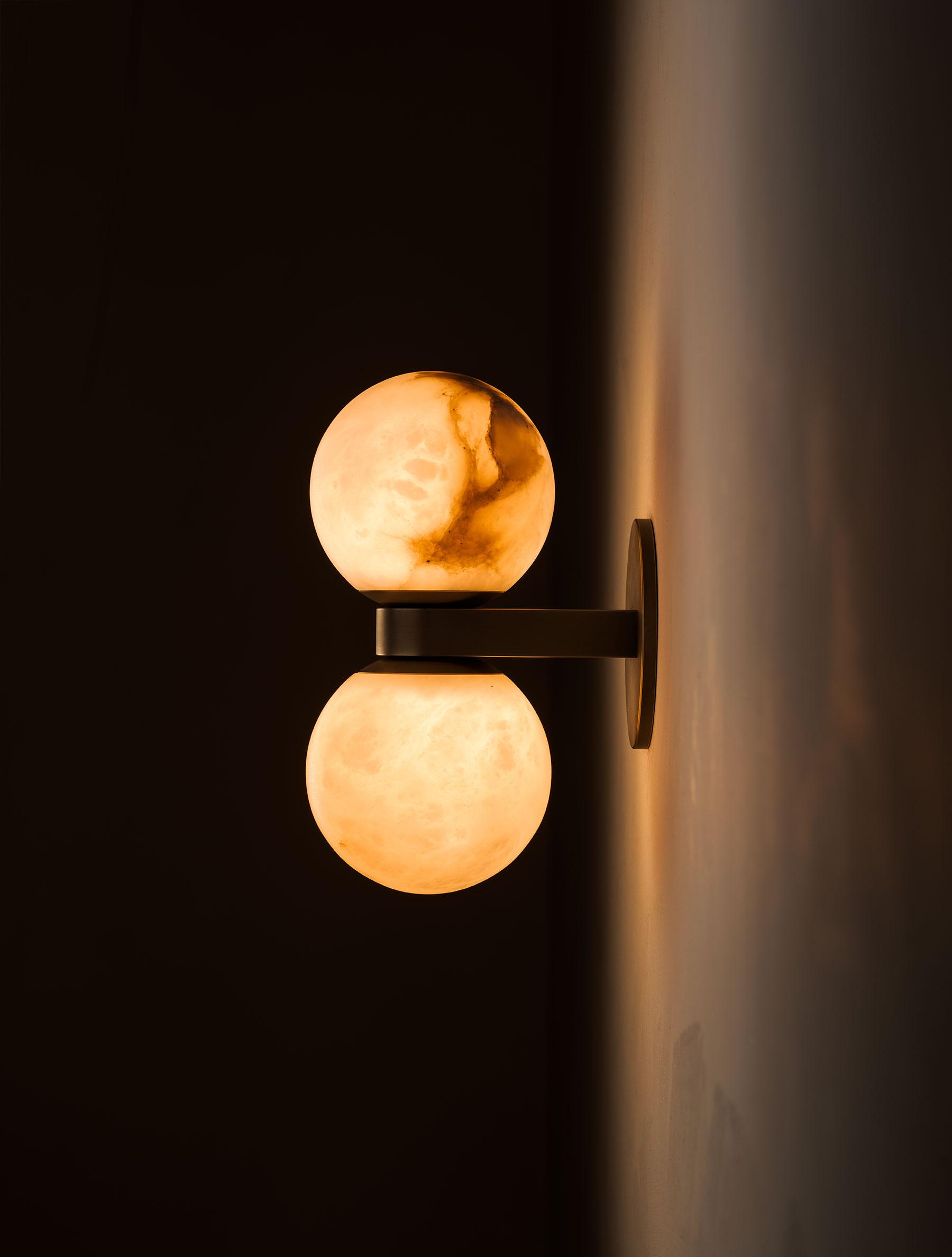 The Clérachic Lush Twin Globe Wall Sconce seen from the side and lit, warm light washing the wall behind its bronze backplate.