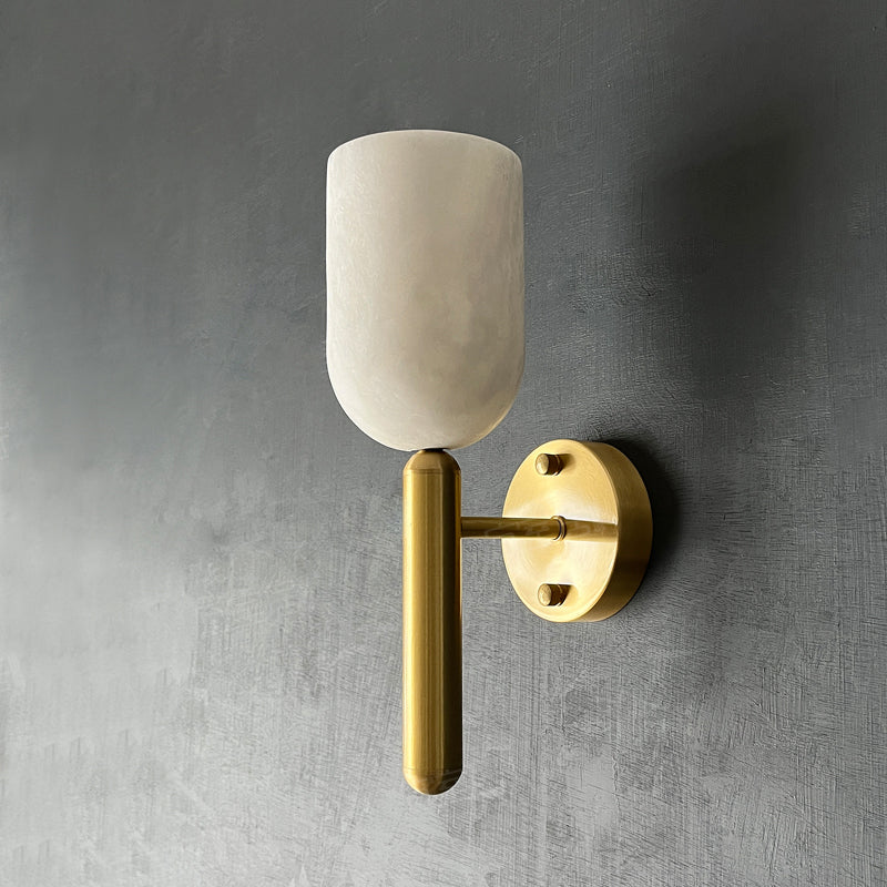 Clérachic Lyphaia wall sconce mounted unlit on a grey plaster wall, the matte alabaster cup sitting above its round brass backplate.