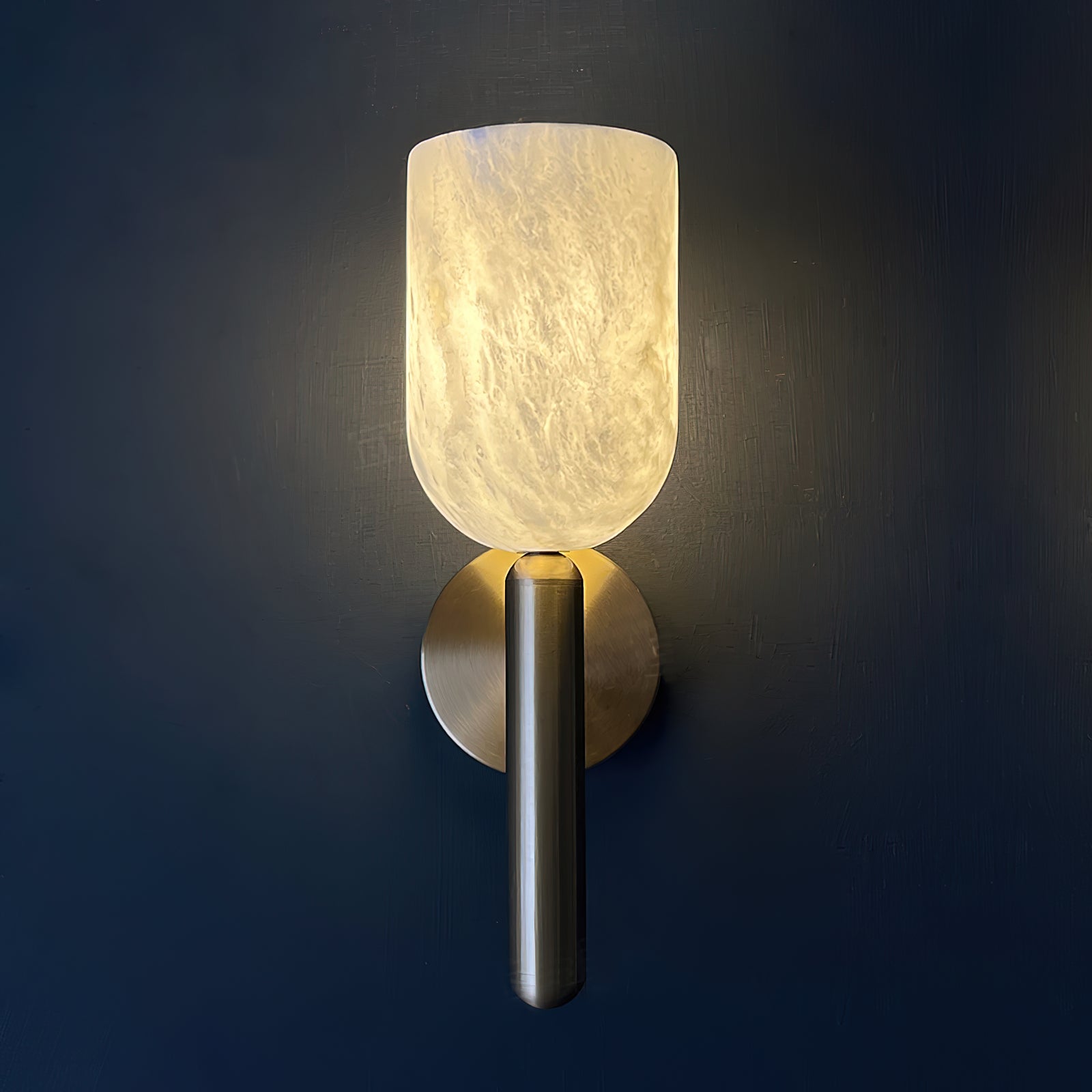 Clérachic Lyphaia sconce lit against a deep navy wall, the alabaster cup turning amber as the veining surfaces from inside the stone.