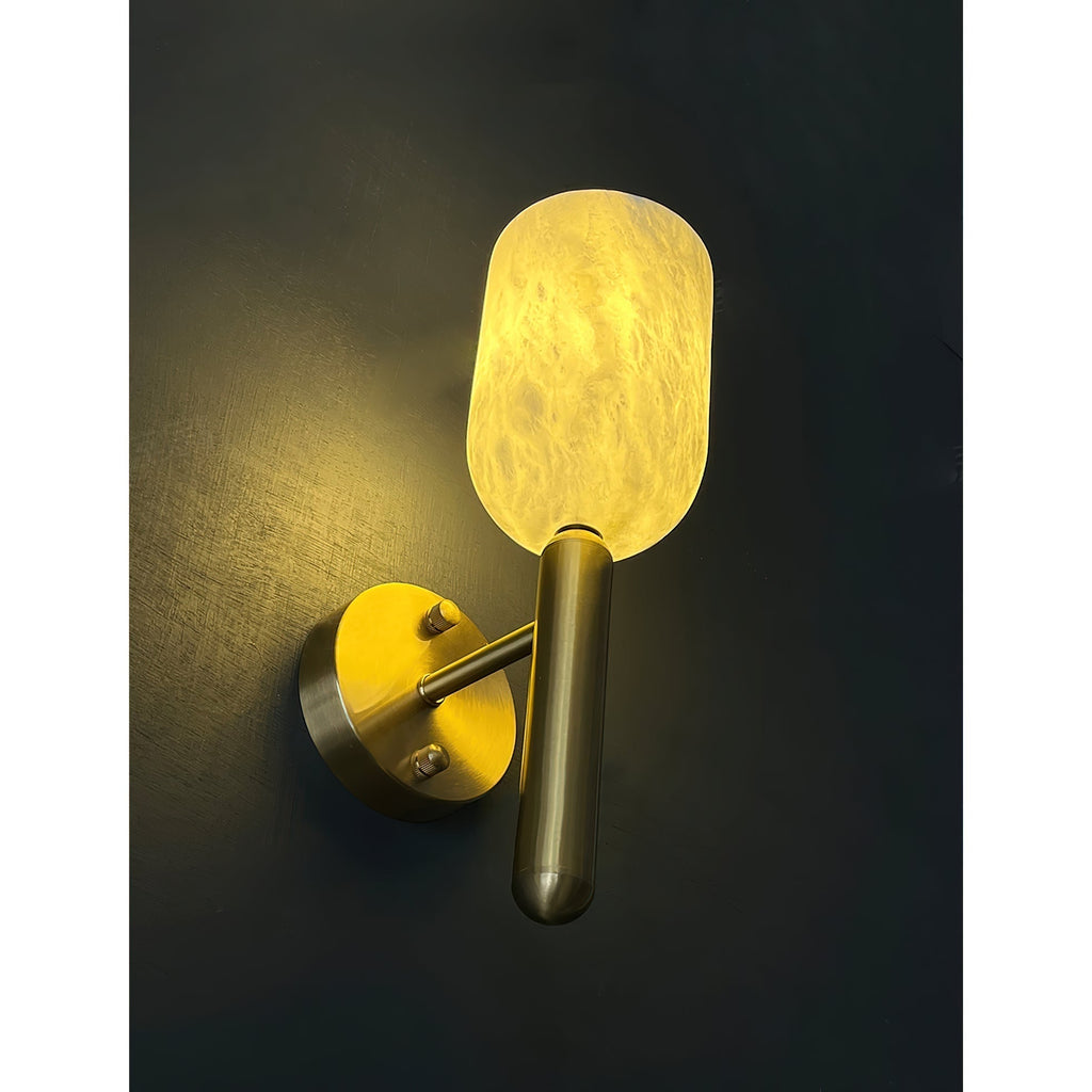 Angled view of the lit Clérachic Lyphaia sconce on a near-black wall, the brass arm and knurled backplate screws catching the glow.