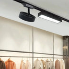 Clérachic black round track light head and a black linear track light on a ceiling rail above a rail of coats in a shop.