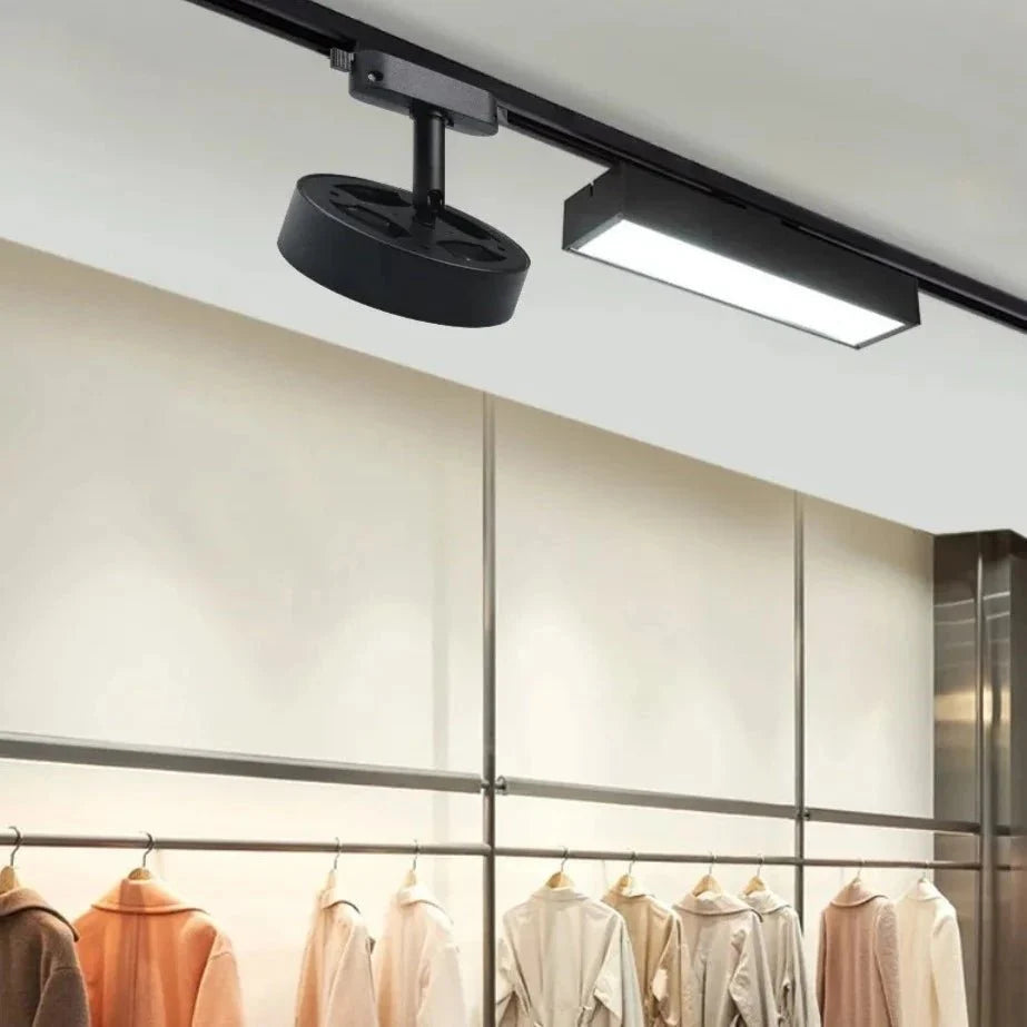 Clérachic black round track light head and a black linear track light on a ceiling rail above a rail of coats in a shop.