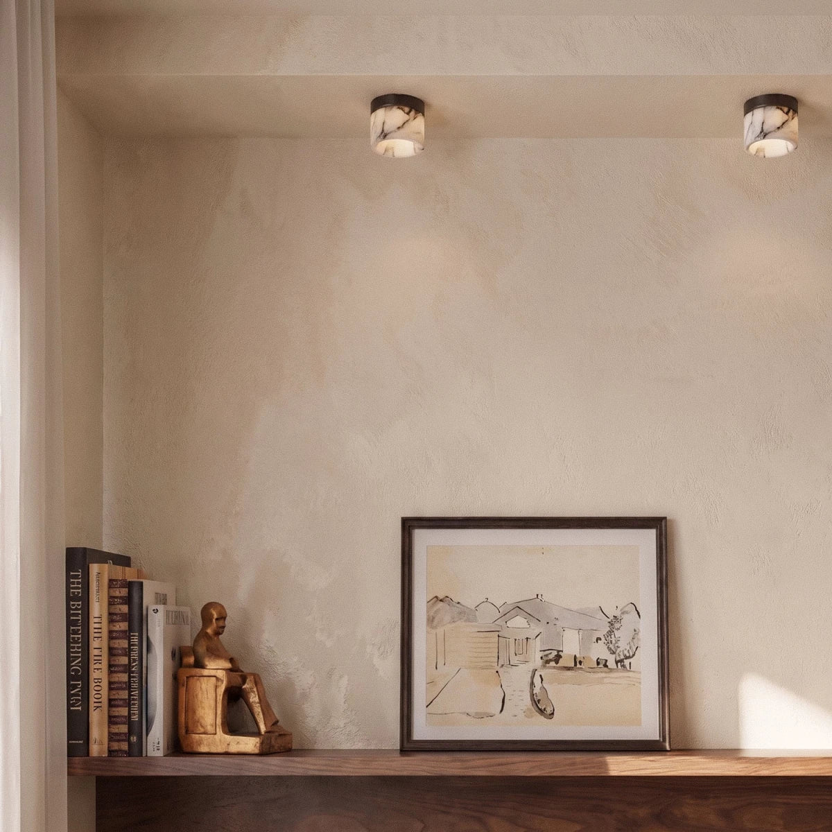 Two Clérachic marble drum ceiling lights above a shelf holding books, a brass figure and a framed ink drawing.