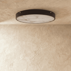 Clérachic flush-mount ceiling light with a veined marble diffuser in a slim dark metal rim, shown on a limewash ceiling.