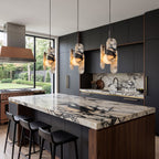 Three Clérachic marble pendants hang over a marble-topped island with a walnut base and black bar stools, in a dark kitchen.