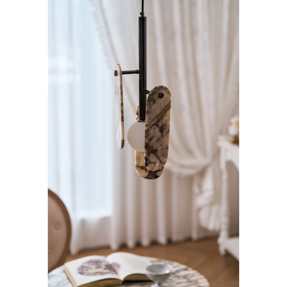 In a cream marble version with violet and amber veining, this Clérachic pendant hangs before a sheer curtain, with an open book on a table below.