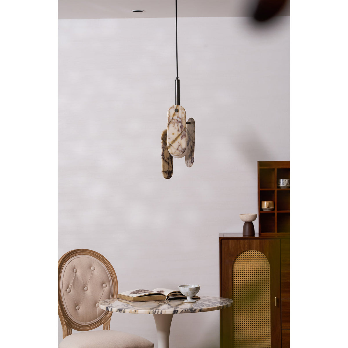 A small Clérachic marble pendant hangs above a round marble-topped table and a tufted chair, beside a rattan-fronted cabinet.
