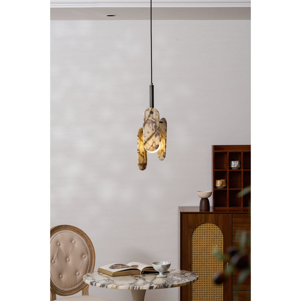 A Clérachic pendant with cream marble panels glows above a marble-topped table, a book and a tufted chair beside a wooden cabinet.