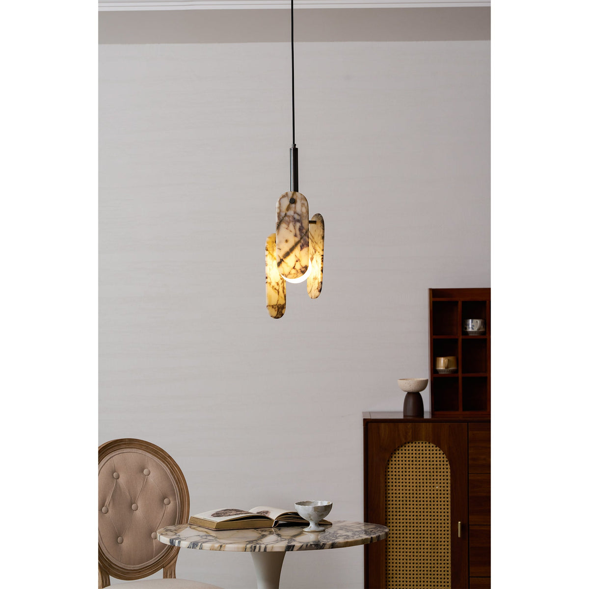 A Clérachic marble pendant glows amber above a round marble table, with an open book, a small cup and a wooden cabinet beside it.