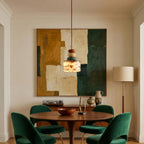 Small Clérachic pendant hung over a round walnut dining table with green velvet chairs, in front of a large abstract painting.