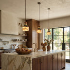 A row of three Clérachic pendants glowing above a marble-topped kitchen island, with wooden cabinets and tall black-framed windows beyond.