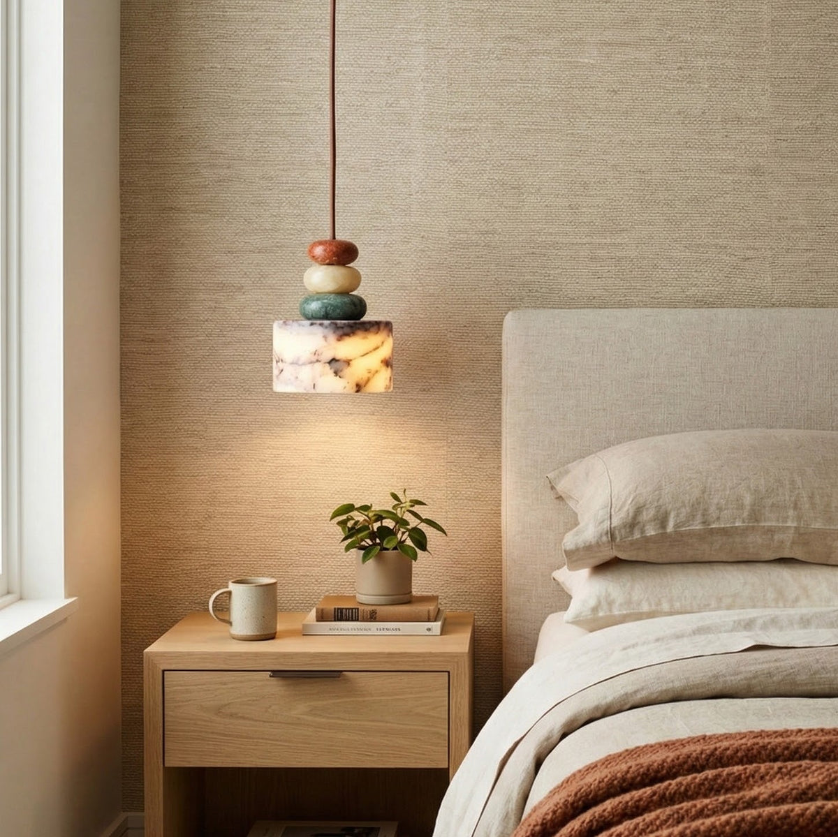 Clérachic pendant hung beside a linen-headed bed, lighting a pale wood nightstand with a plant, a mug and a book.