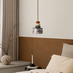Above a bed with cream pillows, a Clérachic pendant hangs against a white wall, with a stone vase of branches and a candle nearby.