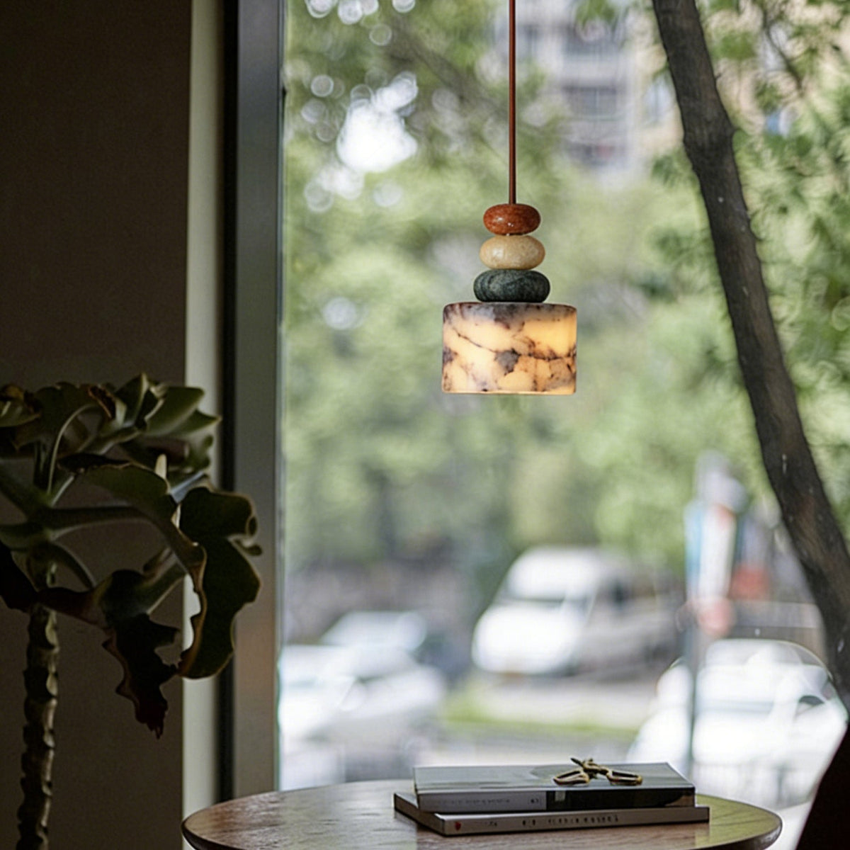 A Clérachic pendant glowing in front of a large window with trees outside, above a round table holding a stack of books.