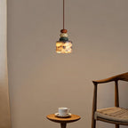 Distant view of a lit Clérachic pendant on a taupe wall, with a wooden armchair and a small round table with a cup beside it.