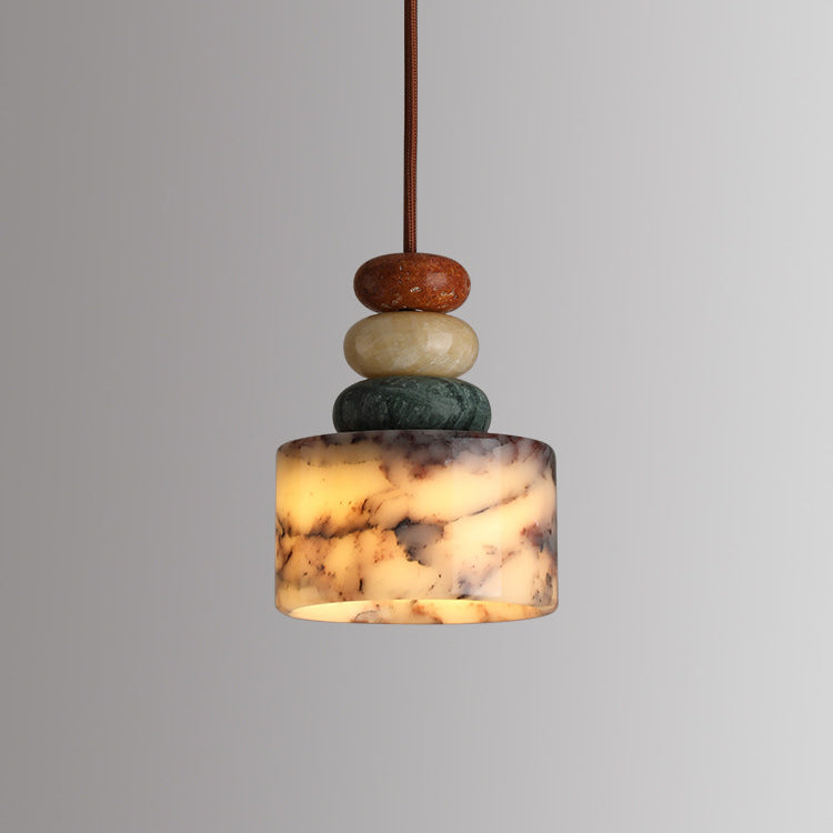 Lit Clérachic pendant on a grey backdrop, the marble drum glowing warm from within while the beads above stay rust, cream and green.