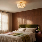 Clérachic stone-slab ceiling light above a bedroom with a walnut panelled wall, olive velvet headboard and linen bedding.