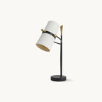 This Clérachic table lamp on a plain white backdrop, with a white drum shade tilted on a black stem with gold accents and a round black base.