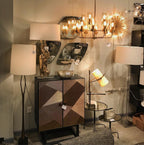 Showroom corner with a Clérachic table lamp, its shade tilted and lit, beside a patterned wood cabinet and other lamps under a gold chandelier.