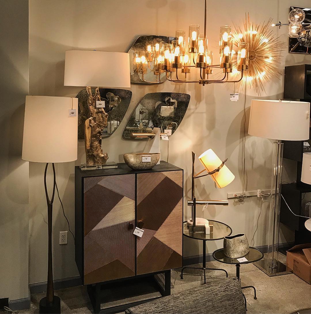 Showroom corner with a Clérachic table lamp, its shade tilted and lit, beside a patterned wood cabinet and other lamps under a gold chandelier.