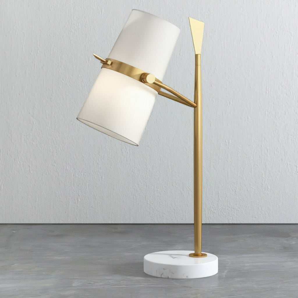 A Clérachic table lamp with a gold stem and white marble base, its white shade angled upward against a pale grey wall.