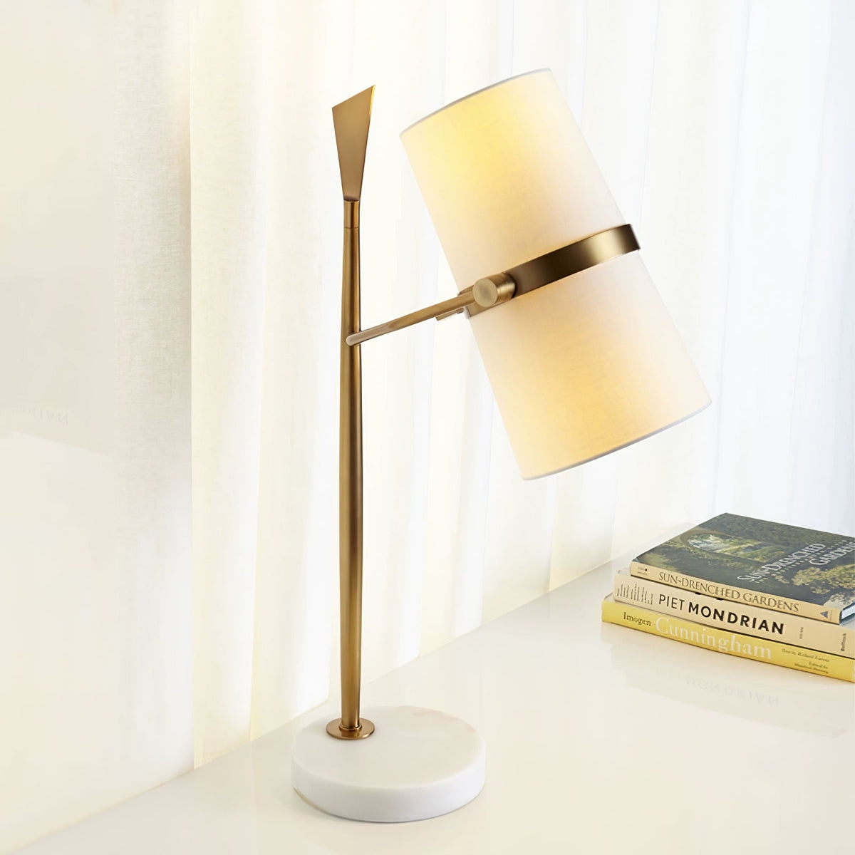 Warm light glows through the white shade of this Clérachic table lamp, which has a gold stem and white marble base, on a desk beside art books.
