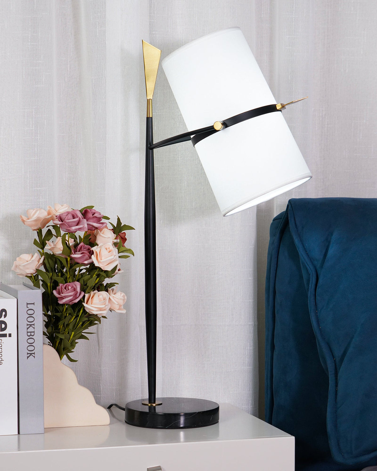 Black-stemmed Clérachic table lamp with a black marble base on a white nightstand, its white shade angled beside pink roses and a blue throw.