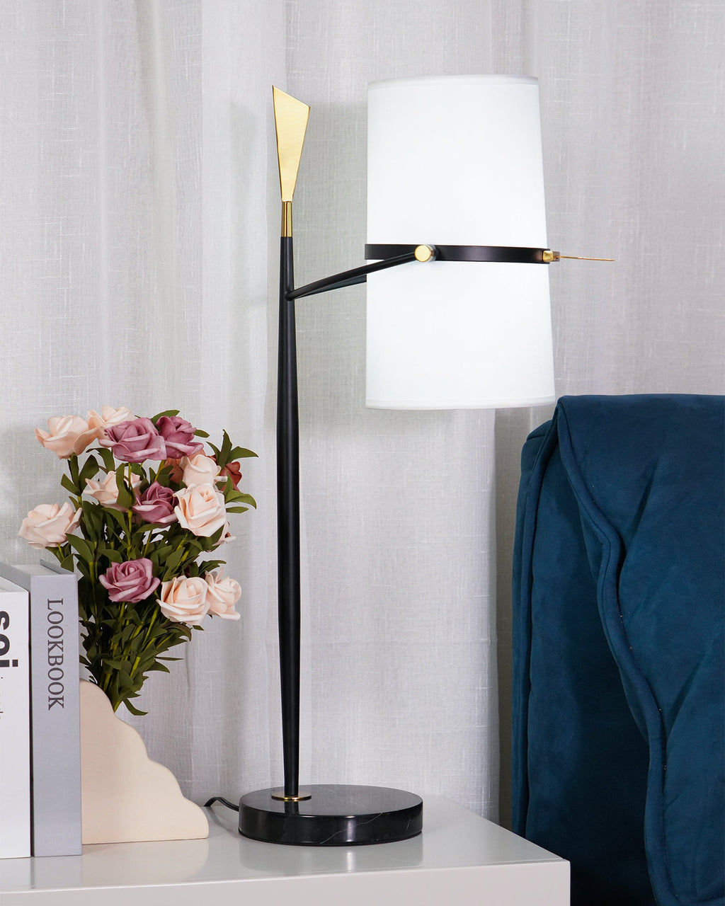 Upright white shade on this Clérachic lamp, with a black stem, gold tip and black base, on a white nightstand beside roses in a vase.