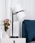 This Clérachic table lamp with its shade tipped sharply downward, on a white nightstand with a drawer, a blue throw and pink roses behind.