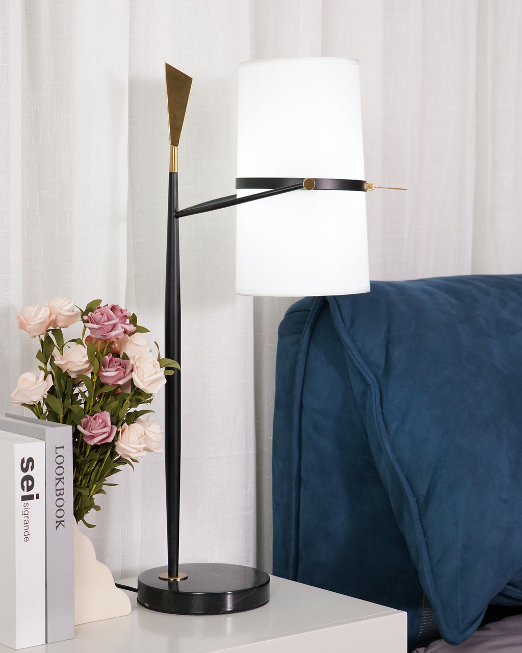 Straight-on view of this Clérachic black table lamp, the white shade held level and the gold tip catching light, beside roses and stacked books.
